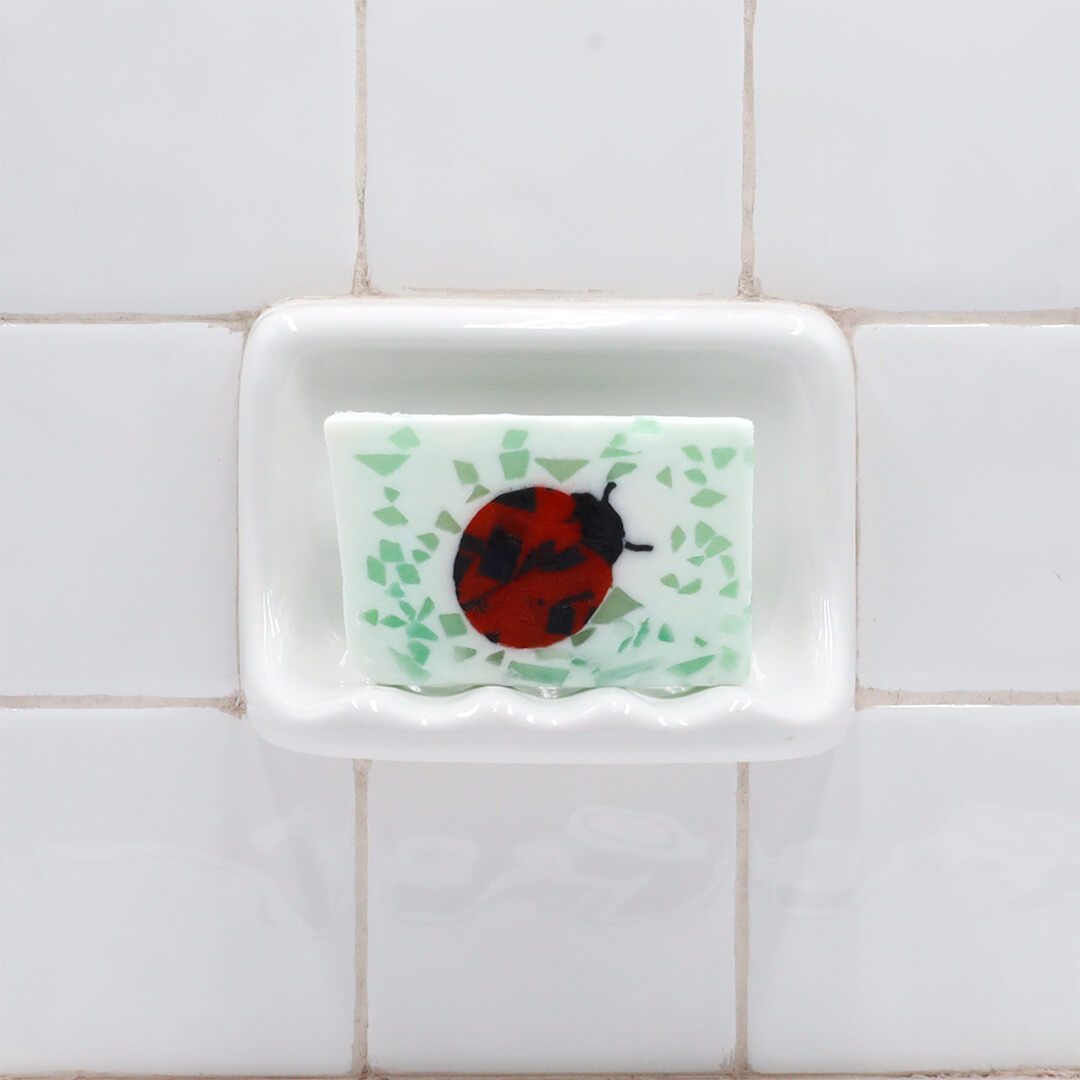 Ladybug Vegetable Glycerin Bar Soap in a white soap dish on a tiled bathroom wall.