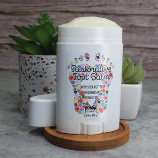 A white foot balm stick labeled 'Restorative Foot Balm' by Primal Elements with a floral foot design, shown on a wooden coaster with its cap removed. The product is styled with two small potted succulents against a rustic wood plank background.