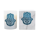 A white glass candle jar with a blue Hamsa hand cutout design, shown next to its white marble-patterned packaging box featuring the same Hamsa design and the text 'ICON CANDLE'.