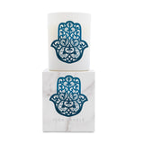 A white candle in a cylindrical holder stacked on top of its square marble-patterned box, both featuring a blue hamsa hand design with intricate cutout details. The box is printed with the brand name 'ICON CANDLE'.