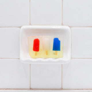 Juice Pops Vegetable Glycerin Bar Soap in a white soap dish on a tiled bathroom wall.