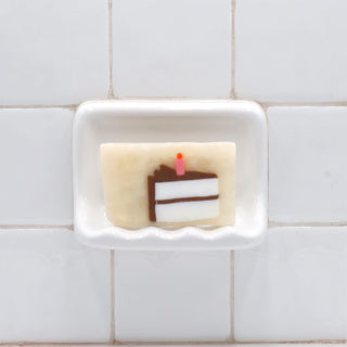 Birthday Cake Vegetable Glycerin Bar Soap in a white soap dish on a tiled bathroom wall.