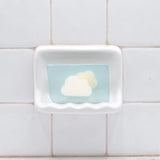 Clouds Vegetable Glycerin Bar Soap in a white soap dish on a tiled bathroom wall.