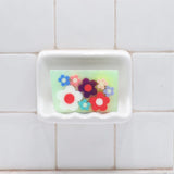 Flowerchild Vegetable Glycerin Bar Soap in a white soap dish on a tiled bathroom wall.