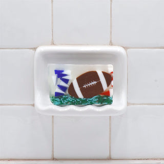 Football Vegetable Glycerin Bar Soap in a white soap dish on a tiled bathroom wall.