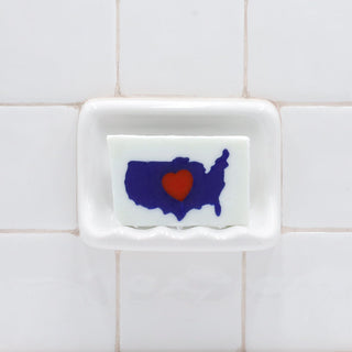 I Heart USA Vegetable Glycerin Bar Soap in a white soap dish on a tiled bathroom wall.