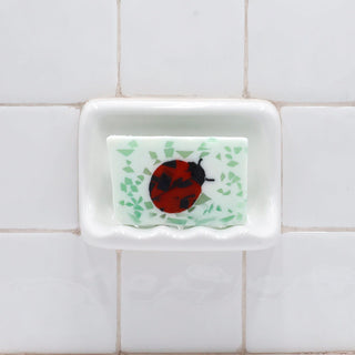 Ladybug Vegetable Glycerin Bar Soap in a white soap dish on a tiled bathroom wall.