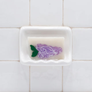 Lilac Vegetable Glycerin Bar Soap in a white soap dish on a tiled bathroom wall.