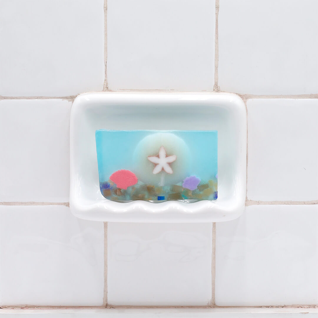 Sand Dollar Vegetable Glycerin Bar Soap in a white soap dish on a tiled bathroom wall.