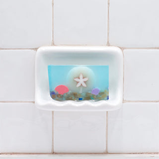 Sand Dollar Vegetable Glycerin Bar Soap in a white soap dish on a tiled bathroom wall.