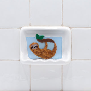 Swinging Sloth Vegetable Glycerin Bar Soap in a white soap dish on a tiled bathroom wall.